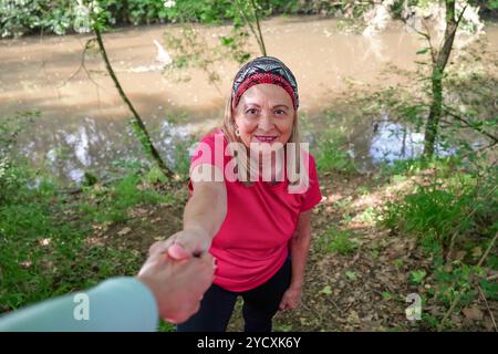 Una donna matura con una camicia rosa brillante e un velo sorride mentre si estende la mano a un'altra persona, accanto a un fiume sereno circondato da una lussureggiante greene Foto Stock