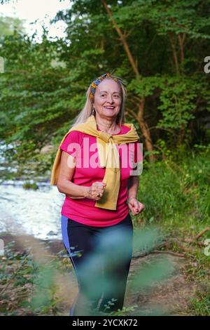 Donna anziana che sorride mentre fa jogging vicino a un ruscello in un parco forestale, vestita con un vivace abbigliamento sportivo e adornata con un archetto colorato Foto Stock