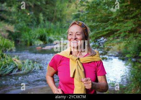 Una donna anziana gioiosa con una vivace cima rosa e una felpa gialla con cappuccio fa una piacevole passeggiata lungo un fiume, circondato da una vegetazione lussureggiante Foto Stock