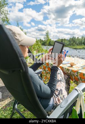 Immortalata in un tranquillo paesaggio svedese, una donna si rilassa vicino a un fiume, profondamente impegnata nella lettura di un eBook su un dispositivo portatile. Foto Stock