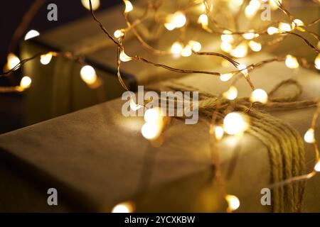 Un primo piano di un regalo di Natale avvolto in carta marrone, legato con spago e circondato da calde luci a corda, che creano una sacra festosa e accogliente Foto Stock