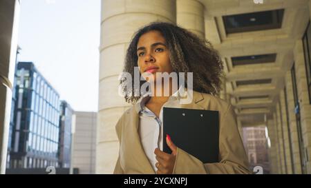 Donna d'affari sicura che cammina in città con gli appunti ed elegante. Sfondo urbano di grattacieli. Rappresenta un leader moderno in vari settori. Riflette la diversità delle donne nella forza lavoro Foto Stock