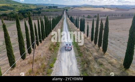 Navigando lungo una tranquilla strada di ghiaia fiancheggiata da alti cipressi, il pittoresco paesaggio toscano si dispiega sotto un cielo limpido, mostrando la tranquillità della vita rurale in Italia. Foto Stock