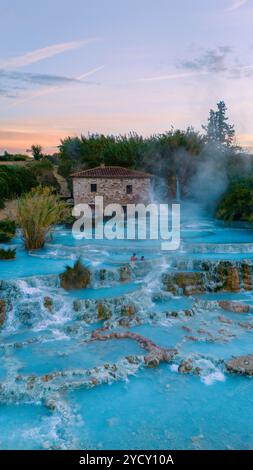 I visitatori si rilassano nelle calde e ricche di minerali piscine termali di Saturnia, circondate da vegetazione lussureggiante e nebbia morbida al tramonto, creando una tranquilla fuga nel pittoresco paesaggio italiano della Toscana Foto Stock