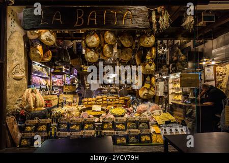 La gastronomia "La Baita" espone prosciutto stagionato all'aria, una selezione di formaggi, pane fresco e confezioni di tagliatelle nella sua vetrina. Un dipendente serve al bancone. Via Pescherie Vecchie, chiamata così in ricordo delle antiche bancarelle di pesce che un tempo si trovavano qui, si trova nel quartiere storico del mercato Quadrilatero. Bologna, Emilia-Romagna, Italia. Foto Stock