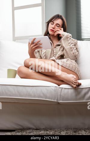 Una giovane donna si siede comodamente su un divano e naviga su un tablet digitale con un'espressione premurosa sul viso. L'atmosfera serena suggerisce una pace Foto Stock