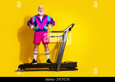 Ritratto completo dell'eccentrico uomo anziano che posa il tapis roulant allenamento in spazi vuoti abbigliamento sportivo retrò isolato su sfondo di colore giallo Foto Stock