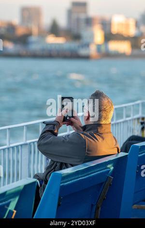 Uomo anziano o di mezza età che utilizza uno smartphone per scattare una foto di Portsmouth mentre attraversa il solent con un servizio di traghetto wightlink per l'isola di wight. Foto Stock