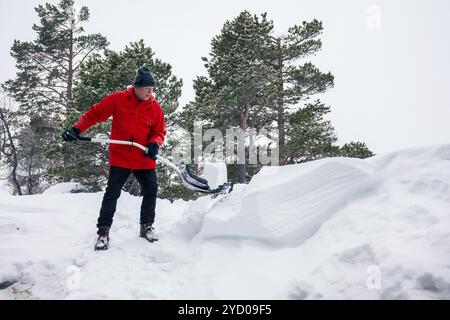 Uomo in giacca rossa che lavora duramente per sparare neve spessa, lavoro fisico, conseguenze delle nevicate, stile di vita rurale, attività invernali Foto Stock