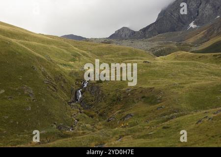 una tranquilla valle di montagna, dove una piccola cascata scende giù da affioramenti rocciosi, che scorre dolcemente attraverso le lussureggianti colline verdi Foto Stock