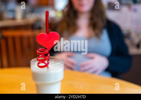 Una vista selettiva creativa di fuoco di un frullato sano spesso imballato con le sostanze nutrienti e le vitamine, servito con la paglia rossa di cuore alle donne pesantemente incinte Foto Stock