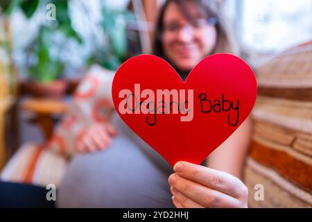 Una vista selettiva messa a fuoco di una donna gravida che tiene un segno di cuore con il messaggio bambino organico, nel terzo trimestre che si prepara per la nascita naturale Foto Stock