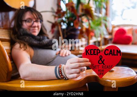 Una vista selettiva di fuoco di una donna gravida che tiene un segno rosso del cuore che dice mantenere la calma ed ottenere pronto per la vita, vicino al lavoro a San Valentino Foto Stock