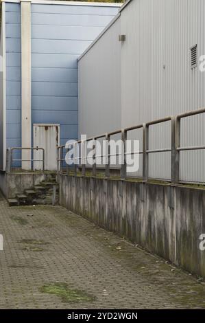 Pareti grigie in cemento fiancheggiano uno stretto corridoio coperto di muschio e foglie morte che conduce a una porta arrugginita Foto Stock