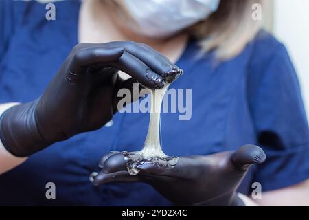 Mani femminili in guanti neri che contengono un recipiente di pasta per shugar pronto per la procedura di epilazione dello zucchero. Trattamenti spa. Beaut femminile Foto Stock