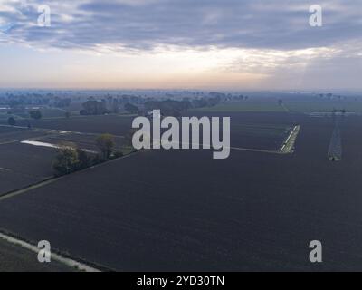 Vista aerea di un pilone elettrico in un campo coltivato, con una foresta e un piccolo villaggio sullo sfondo al tramonto Foto Stock