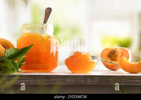 Vaso e recipiente con marmellata di pesca, frutta e fette con foglie su un tavolo di legno e cucina sullo sfondo. Vista frontale. Foto Stock