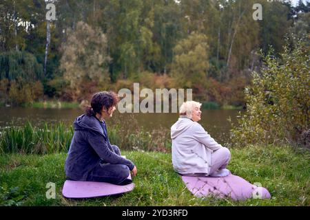 una donna di mezza età insegna alla sua vecchia madre esercizi somatici e yoga per mantenere la salute sul fiume in autunno Foto Stock