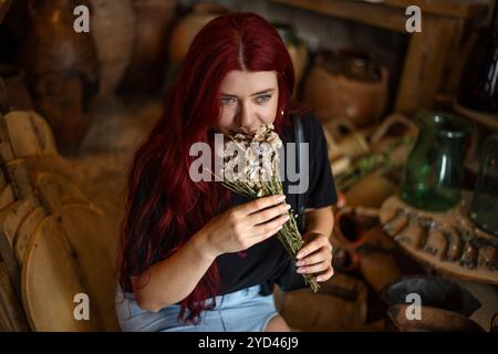 Giovane bella donna seduta in un vecchio laboratorio e odorante di fiori secchi Foto Stock
