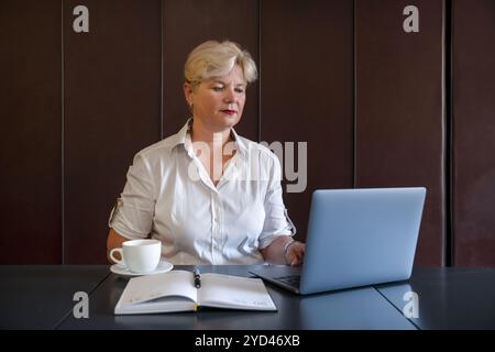Donna di mezza età seduta a un tavolo con una tazza di caffè, un open planner e che guarda lo schermo di un notebook con un bagno composto e professionale Foto Stock
