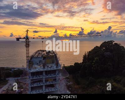 Vista droni di un edificio a più piani in costruzione e di una gru da costruzione sullo sfondo di un bellissimo tramonto sul mare. Concetto di costruzione Foto Stock