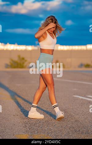 Donna sportiva cammina su una strada tranquilla al tramonto dopo un allenamento, incarnando uno stile di vita sano. Trasuda energia e motivazione nel suo abbigliamento sportivo, godendosi la serata tranquilla Foto Stock