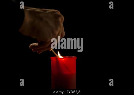 Mano maschile, usando un abbinamento di sicurezza, illuminando una candela rossa del cimitero, su uno sfondo nero, primo piano. Concetto di giorno di tutti i santi. Foto Stock
