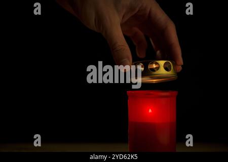 Mano maschile, usando un abbinamento di sicurezza, illuminando una candela rossa del cimitero, su uno sfondo nero, primo piano. Concetto di giorno di tutti i santi. Foto Stock