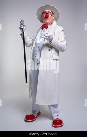 Un clown anziano con un orologio antico su una catena in mano. Vestito con un cappotto bianco, un cappello e un grande clown in pelle verniciata rossa Foto Stock