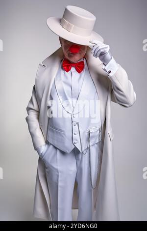Un grosso clown mafioso anziano con un cappotto e un cappello bianco. Mano destra nella tasca, mano sinistra regola il cappello. Foto Stock