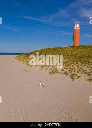 Una coppia si trova fianco a fianco su una spiaggia sabbiosa vicino ad un faro rosso e bianco, bagnata dalla calda luce del tramonto sullo sfondo Foto Stock