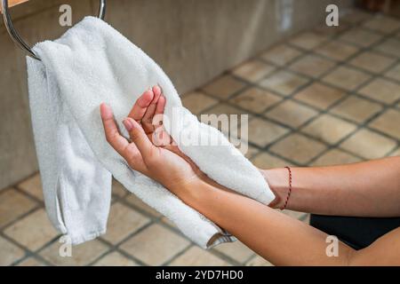 Un primo piano delle mani di una persona che si asciuga con un asciugamano bianco in un bagno. Lo sfondo presenta un portasciugamani in legno e pareti piastrellate, creando Foto Stock