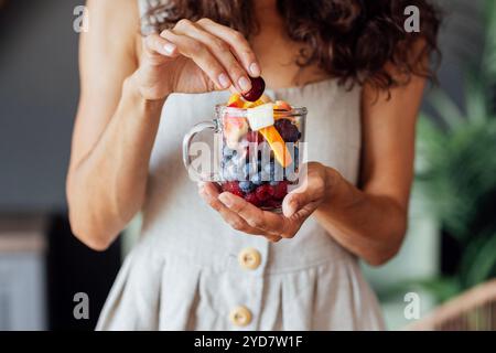 Primo piano di una tazza di vetro con frutta e frutti di bosco in mani femminili ben curate. Una giovane donna in un prendisole sta mangiando ciliegie A. Foto Stock