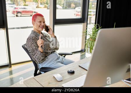 Una donna allegra e calva si impegna in una telefonata vivace mentre si siede nel suo moderno spazio di lavoro. Foto Stock