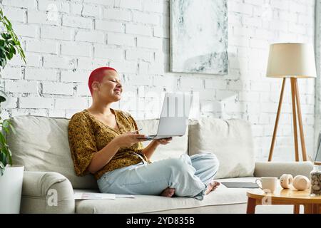 Una donna allegra con una testa calva si rilassa sul divano mentre si impegna con il suo laptop. Foto Stock