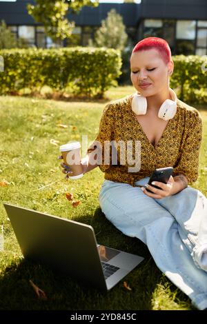 Una donna calva e felice si diverte con un drink caldo e si impegna con il suo notebook mentre è seduta sull'erba. Foto Stock