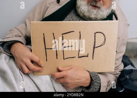 Primo piano ritagliato di un senzatetto anziano irriconoscibile seduto sul freddo marciapiede della città, con un cartello che chiede aiuto. Concetto di povertà e. Foto Stock