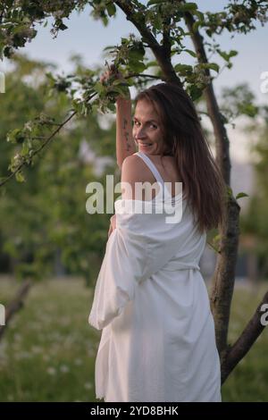 Una donna in abiti bianchi si appoggia contro un albero con le braccia sollevate, sembrando rilassata. Questa immagine cattura l'armonia tra uomo e natura in un Foto Stock