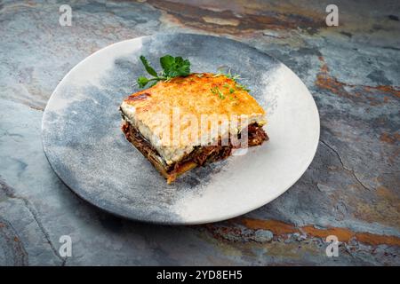 Moussaka greca tradizionale con carne di manzo, melanzane e salsa di besciamella servita come primo piano su un piatto di design nordico su una tavola di pietra Foto Stock