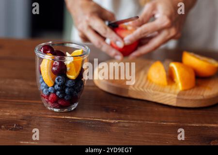 Primo piano di una ciotola di vetro con frutta e frutti di bosco e mani femminili. Una giovane donna sta affettando un'insalata di frutta fresca. Foto Stock