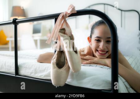Una studentessa di danza femminile guarda le vecchie scarpe da ballo appese alla fine del letto, sorriso felice Foto Stock