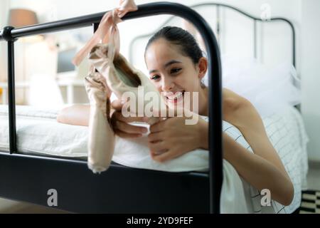 Una studentessa di danza femminile guarda le vecchie scarpe da ballo appese alla fine del letto, sorriso felice Foto Stock