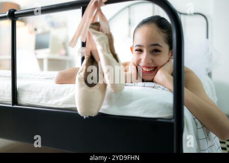 Una studentessa di danza femminile guarda le vecchie scarpe da ballo appese alla fine del letto, sorriso felice Foto Stock