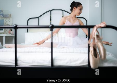Una studentessa di balletto si riscalda a letto e guarda le vecchie scarpe da balletto appese alla fine del letto Foto Stock