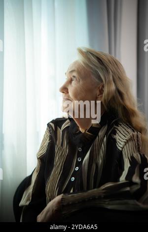 Una vecchia donna solitaria si siede e guarda fuori dalla finestra dove splende il sole del mattino. Mi mancano i miei cari che sono stati insieme per un po' Foto Stock