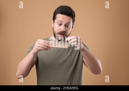 Uomo emotivo che prende i capelli persi dal pettine su sfondo beige. Problema di alopecia Foto Stock