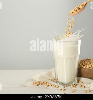 I semi di soia cadono dal cucchiaio di legno in un bicchiere di latte vegetale sul tavolo bianco. Spazio per il testo Foto Stock