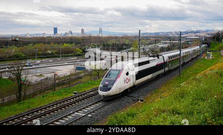 Un treno TGV InOui attraversa una montagna in primavera, sotto un cielo nuvoloso ma limpido, con lo skyline del centro di Lione visibile sullo sfondo. Foto Stock