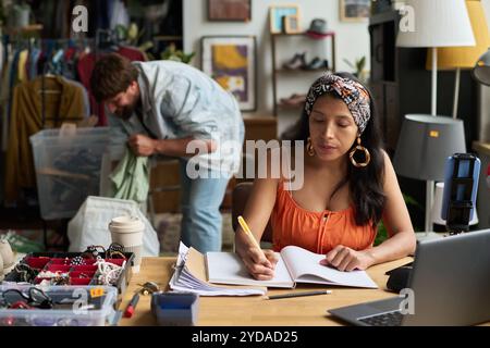 Giovane donna in elegante abbigliamento casual che prende appunti nel copybook mentre organizza il lavoro contro assistente maschile con container Foto Stock