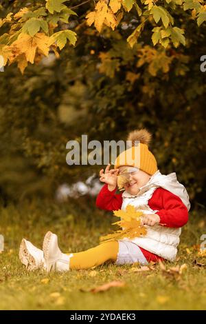 Un bambino sorridente in un abito luminoso siede sull'erba, giocando con gioia con le vibranti foglie autunnali circondate da fogliame colorato in una giornata di sole. Foto Stock
